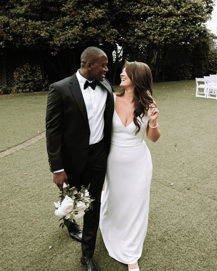 Bride and groom walking on grass, smiling at each other, holding a bouquet.