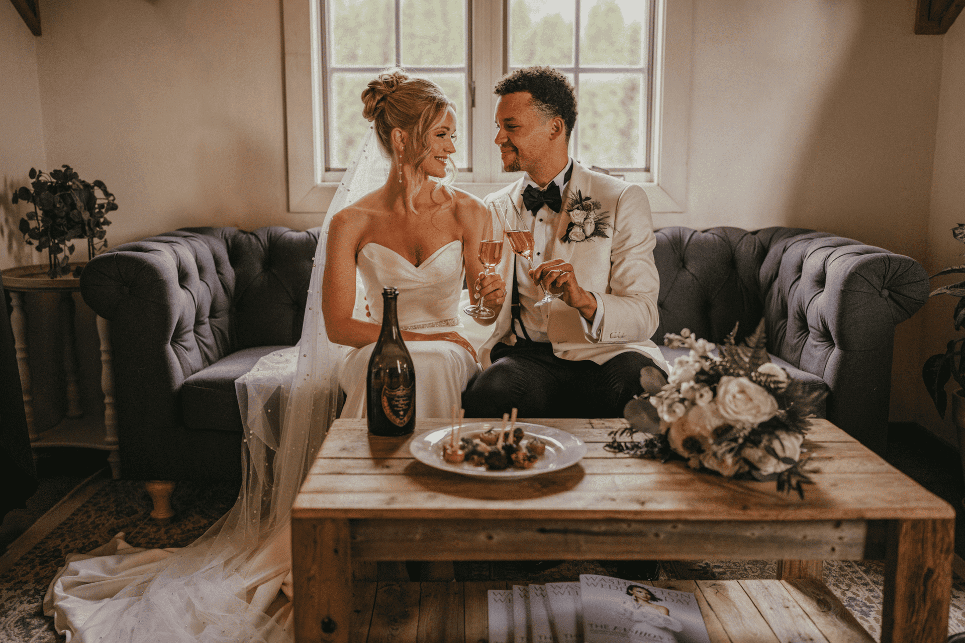 Bride and groom toasting with champagne on a cozy couch, wedding bouquet and decor in foreground.